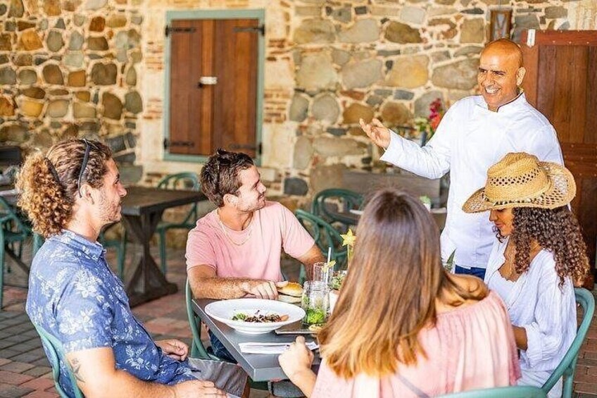 A group of people dining outdoors at a table with a stone wall and wooden door in the background. 2