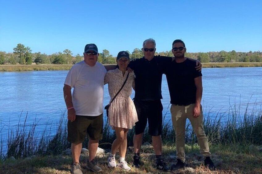 Four people standing by a lake with grass and trees in the background. 4