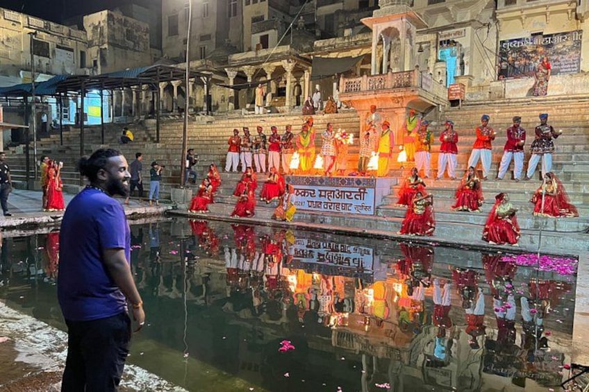 A group of people in traditional attire performing a ritual by a river with a temple in the background. 1