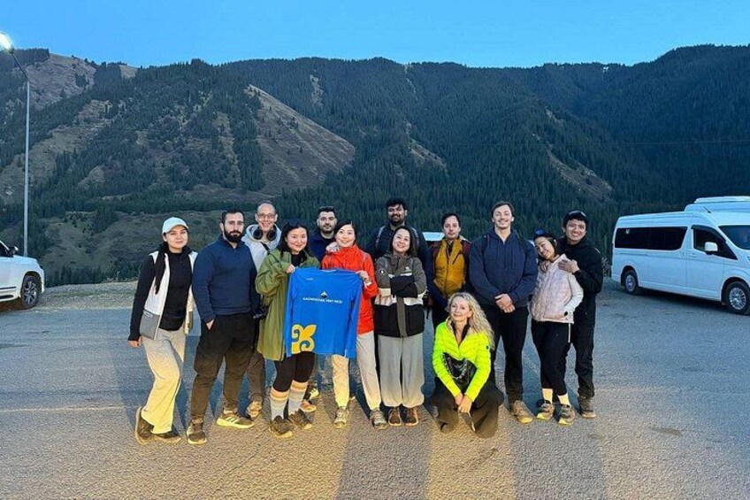 A group of people posing in front of a mountainous landscape with a white van and a car in the background. 2