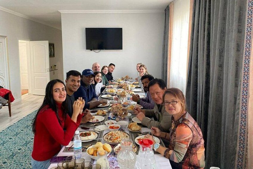 A group of people seated around a long dining table with various dishes, in a room with a television on the wall and a patterned curtain. 2