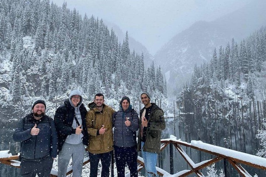 Eine Gruppe von Menschen steht auf einer mit Schnee bedeckten Holzplattform, im Hintergrund schneebedeckte Bäume und Berge. 5