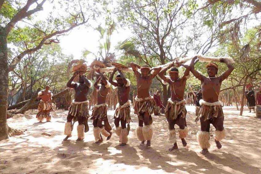 A group of people in traditional attire performing a dance in an outdoor setting with trees and a dirt ground. 5