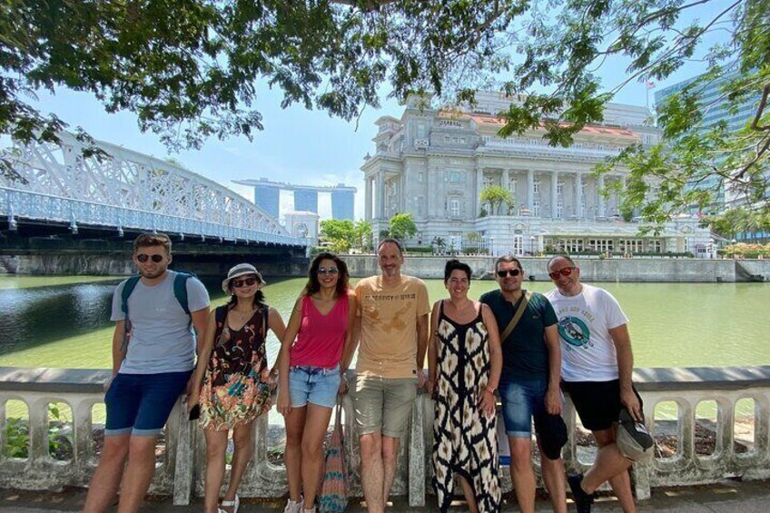 A group of people standing by a river with a bridge and a large building in the background. 1