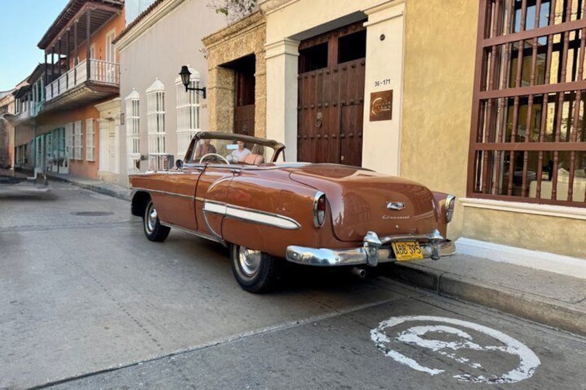 Ein brauner, klassischer Oldtimer-Cabrio steht auf einer Straße, im Hintergrund sind Gebäude und eine 'Coca-Cola'-Werbetafel zu sehen. 2