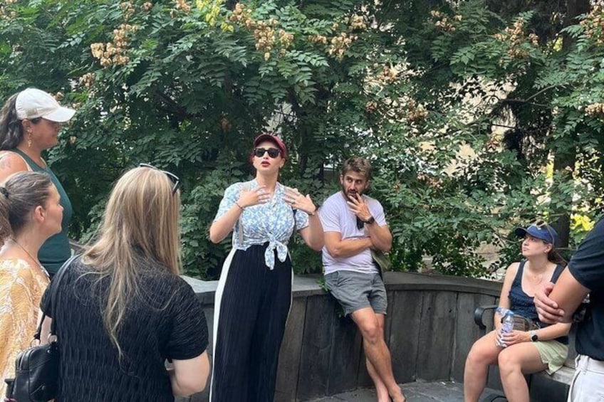 A group of people sitting and standing outdoors, with trees and a stone wall in the background. 4
