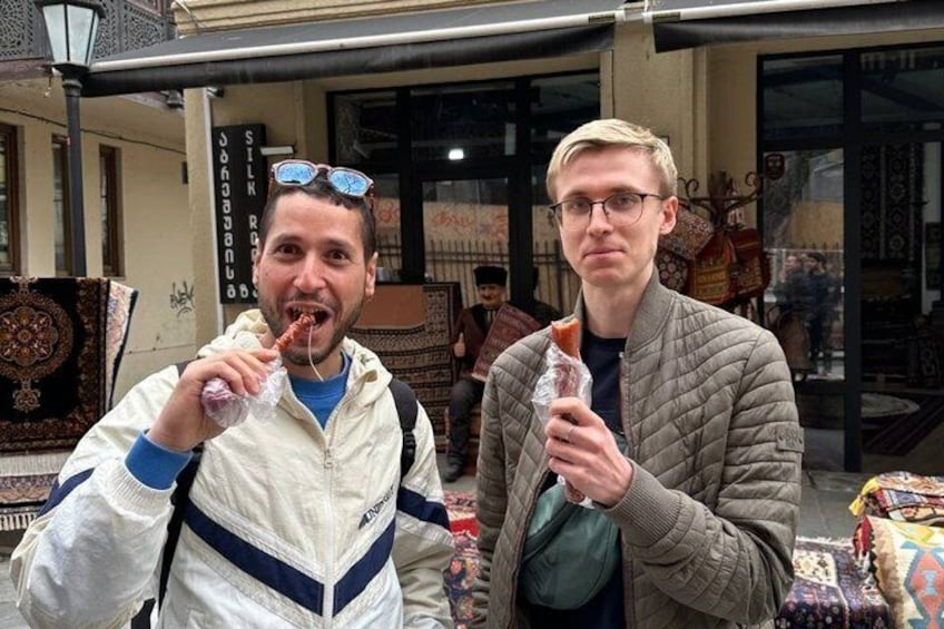 Two people eating food in an outdoor market with patterned rugs and textiles in the background. 2