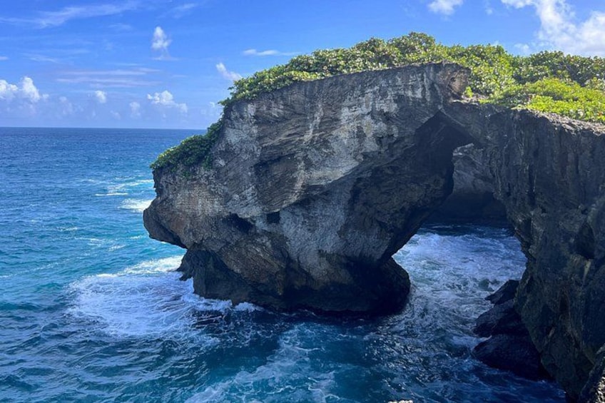 A natural rock arch formation in the ocean with clear blue water and a small island with vegetation on top. 1