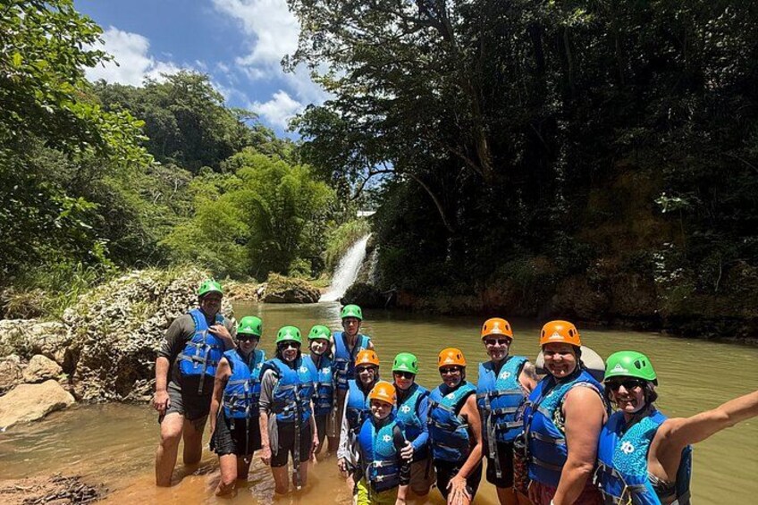 A group of people in blue life vests and helmets, standing by a river with a waterfall in the background. 2