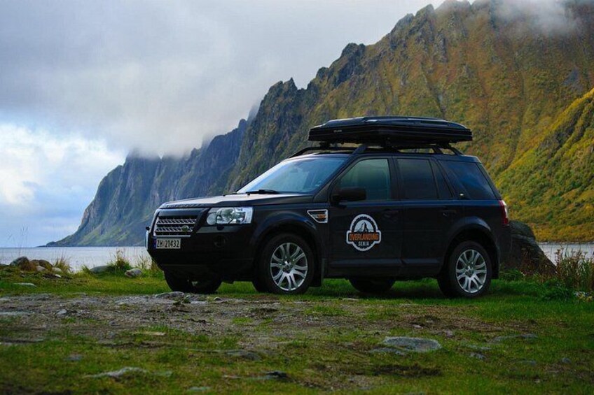 Un SUV negro con portaequipajes estacionado en un césped con montañas y una masa de agua al fondo. 2