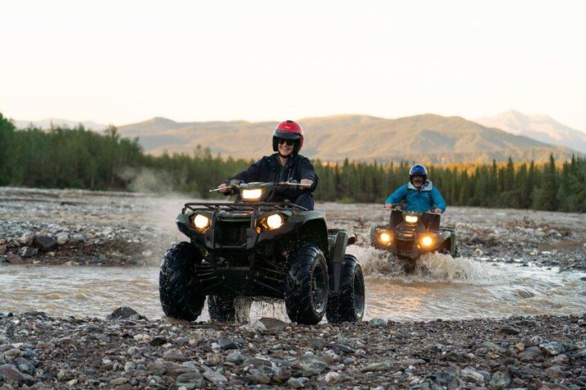 Two people riding ATVs on a rocky terrain with a mountainous background. 3