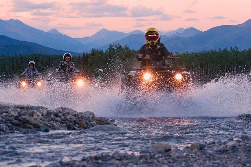 Three people riding all-terrain vehicles on a rocky riverbed with mountains in the background. 4