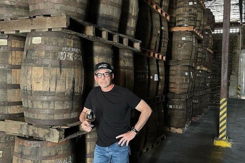 A person standing in a warehouse with wooden barrels stacked around. 4