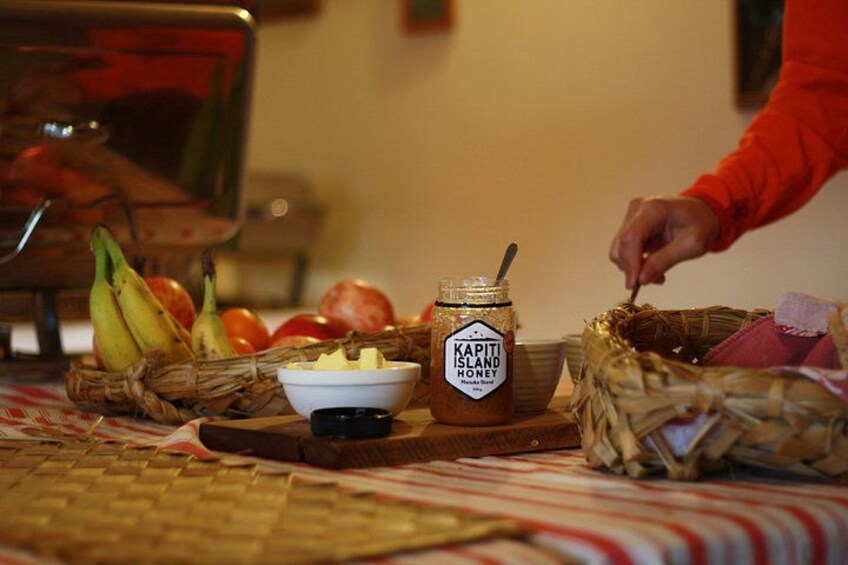 A table with a jar of Kapiti Island honey, a bowl of butter, bananas, apples, and a basket. 4