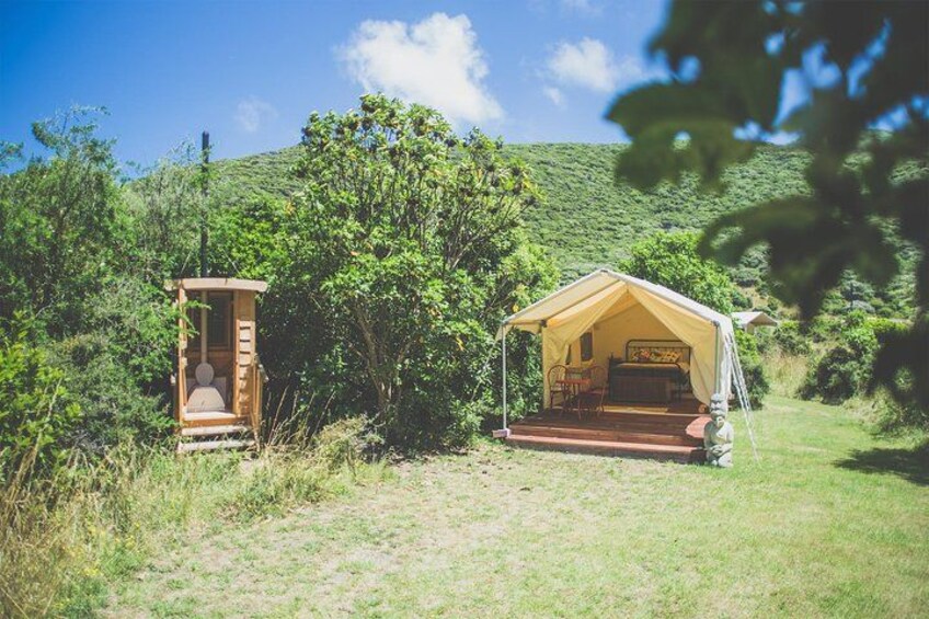 A tented camping area with a small wooden structure and a larger tent set up in a grassy field with trees and hills in the background. 3