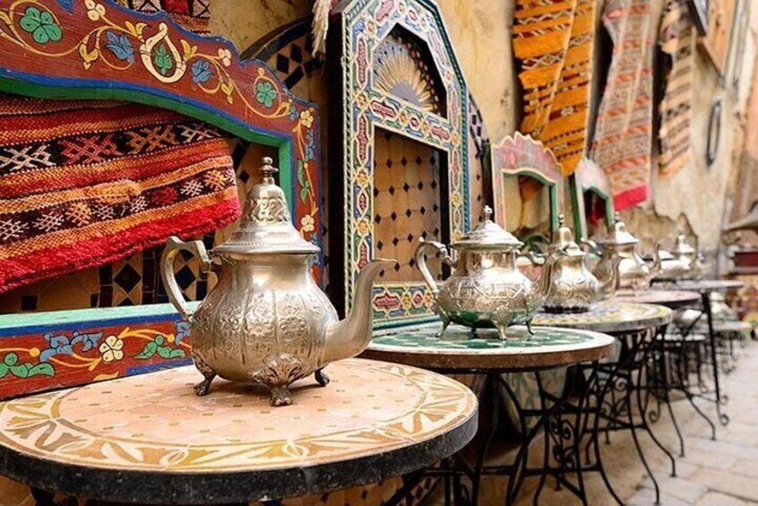 Ornate metal teapots on outdoor tables with colorful textiles and traditional Moroccan architecture in the background. 5