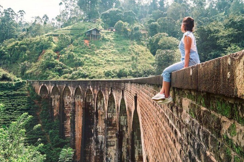 A person sitting on a bridge with a steep drop below, surrounded by lush greenery and hills. 3