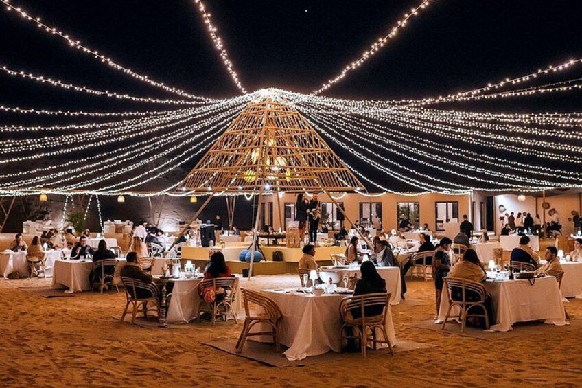An outdoor dining area with tables and chairs, string lights overhead, and a wooden structure in the background. 1