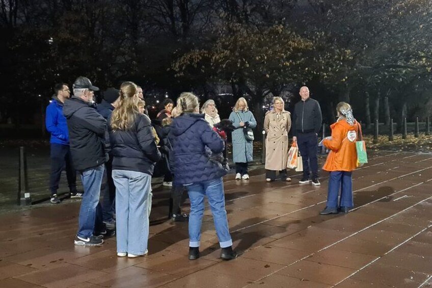 A group of people standing on a wet pavement at night, likely in a park or public space. 2