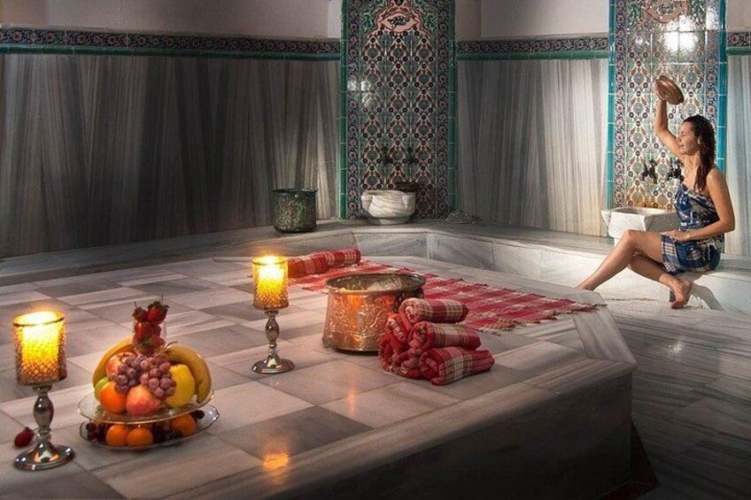 Una habitación con piso de mármol, una mujer sentada en un banco y una mesa con fruta y velas. 4