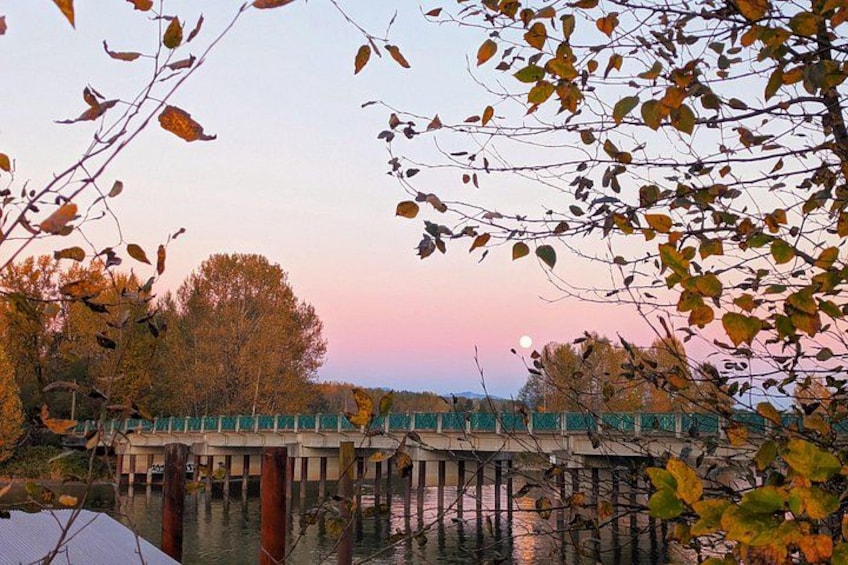 Un puente de madera sobre el agua, rodeado de árboles con hojas otoñales, y una puesta de sol al fondo. 2