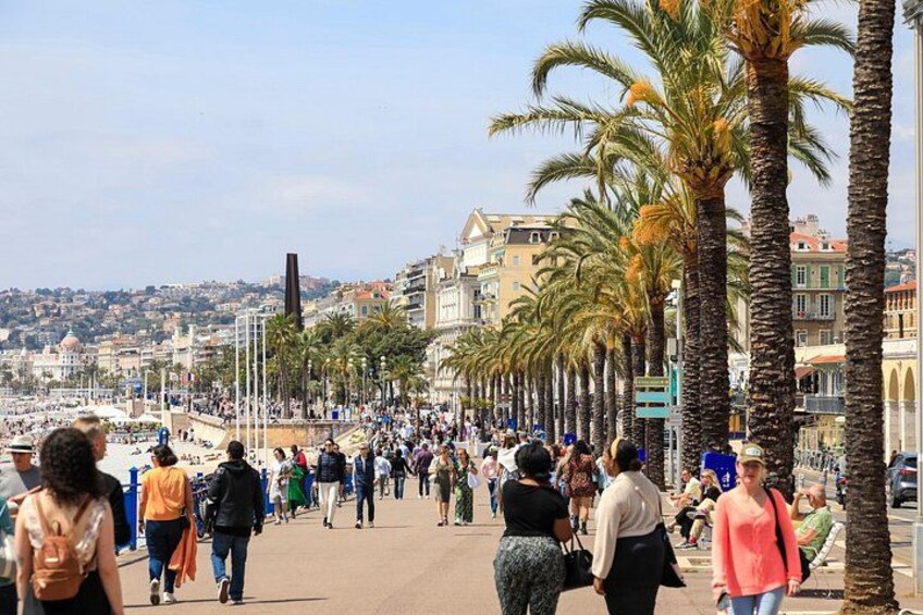 A crowded promenade lined with palm trees and buildings, with people walking and sitting. 4