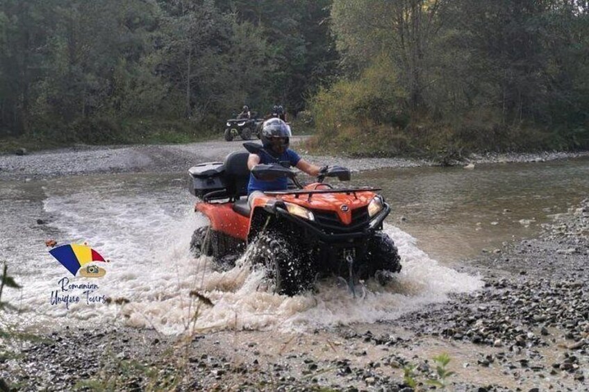 An ATV navigating through a shallow river surrounded by trees. 3