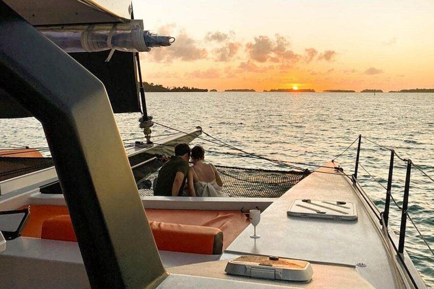 A boat with a person holding a wine glass, another person sitting, and a sunset in the background. 1