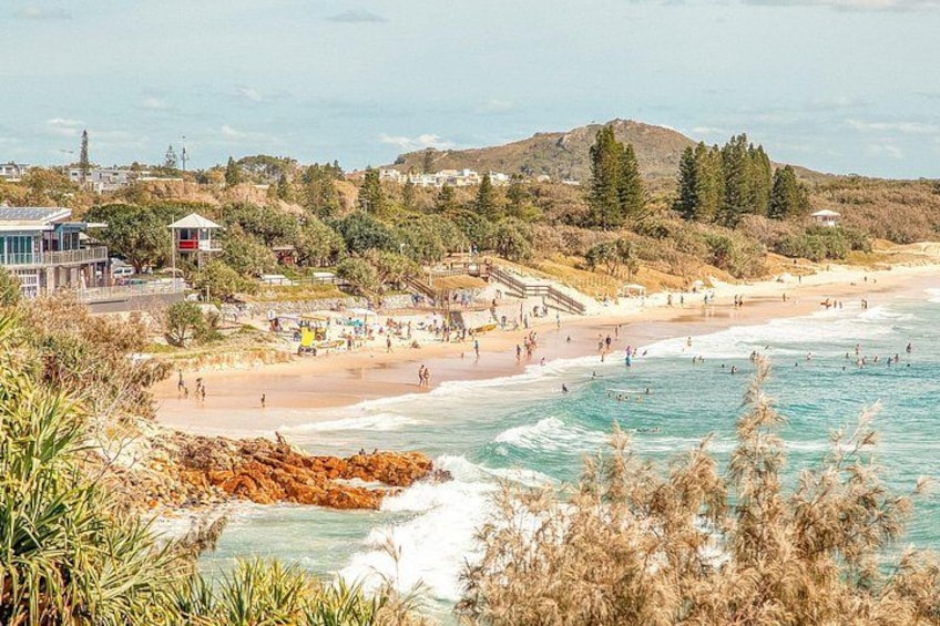 A beach with people swimming and walking, a rocky shoreline, and buildings in the background. 1