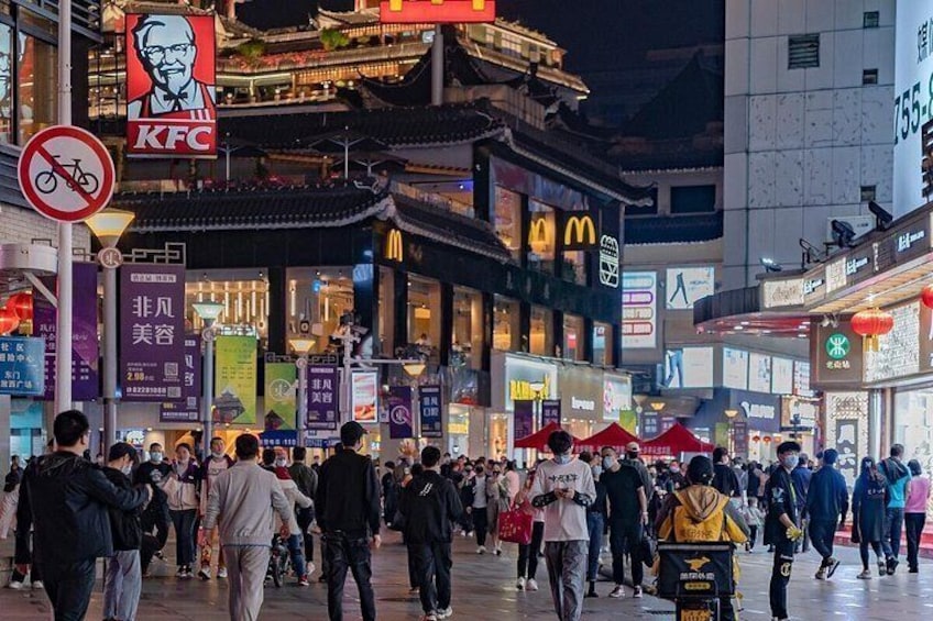Eine belebte Stadtstraße bei Nacht mit beleuchteten Schildern, darunter ein KFC-Logo, und traditioneller Architektur. 3