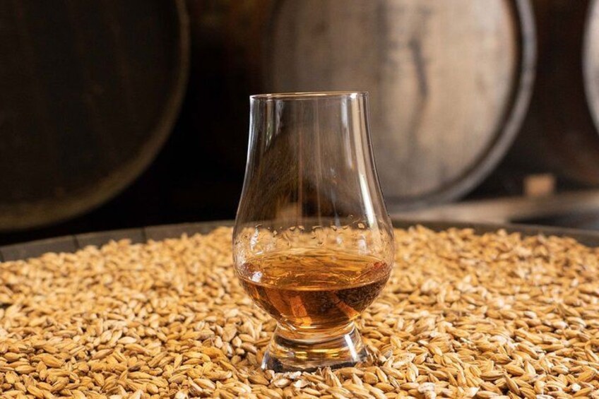 Ett glas med en bärnstensfärgad vätska, troligen whisky, placerat på ett underlag av spannmål, med tunnor i bakgrunden. 2