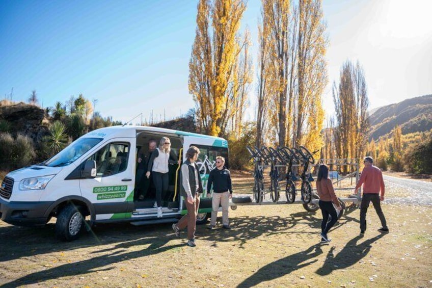 A white van with a green stripe parked on a dirt road, surrounded by autumn trees and a clear sky. 4