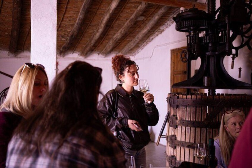 A group of people in a rustic cellar with wooden beams, a large wine press, and bottles on shelves. 2