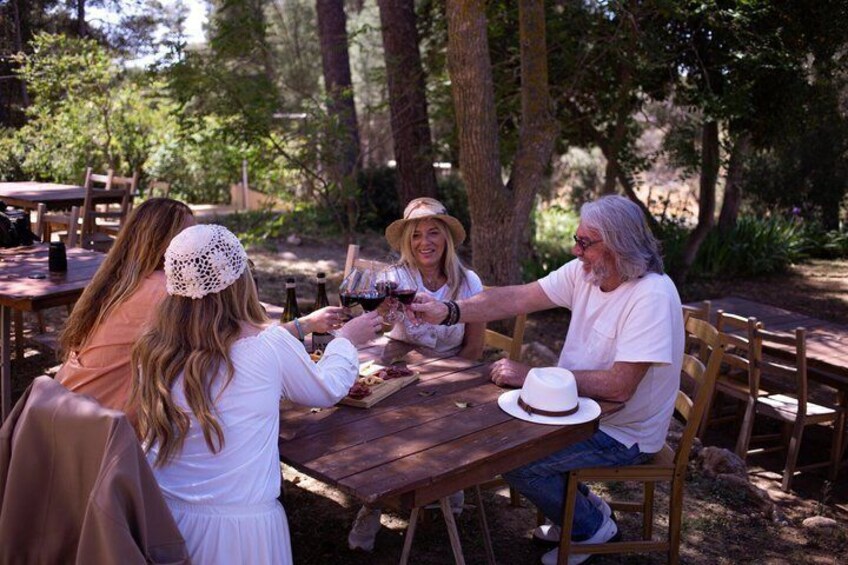 A group of people sitting at a wooden table outdoors, engaged in a meal with wine glasses. 4