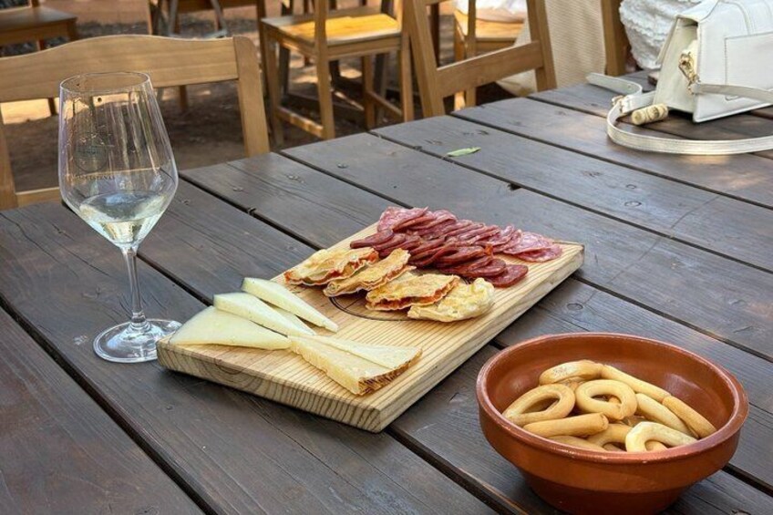 A wooden table with a charcuterie board, a glass of white wine, and a bowl of pretzels. 3