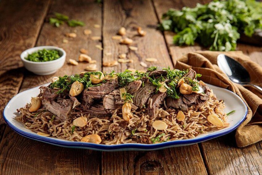 En tallerken med biryani, kød og ris, pyntet med nødder og krydderurter, står på et træbord. 1