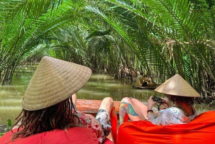 Two people sitting on a boat in a lush, green environment with dense foliage. 1