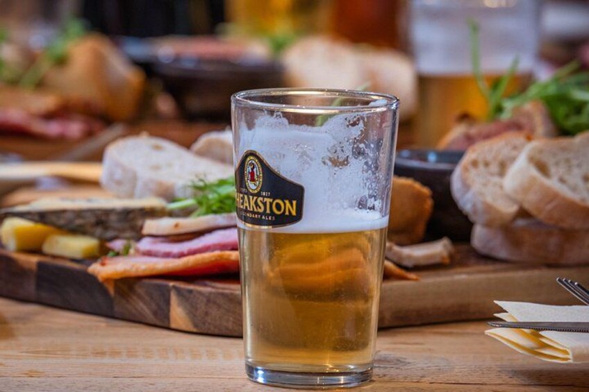 A glass of beer with a label, a wooden board with various meats and bread, and a blurred background of a dining setting. 3