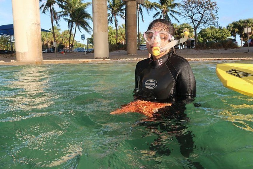 Une personne vêtue d’une combinaison de plongée noire, tenant un poisson dans une piscine bordée de palmiers, avec un kayak jaune en arrière-plan. 4