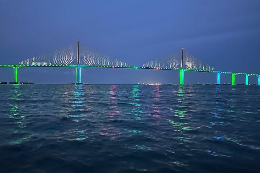 Eine Brücke mit grüner Beleuchtung bei Nacht, deren Spiegelung sich im Wasser zeigt. 1