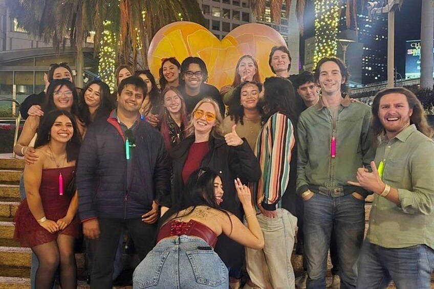 Eine Gruppe von Menschen posiert für ein Foto in einer Stadt, im Hintergrund eine große herzförmige Struktur und Stadtlichter. 3