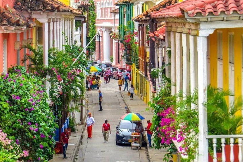 A narrow street lined with colorful buildings, people walking, and parked cars. 1