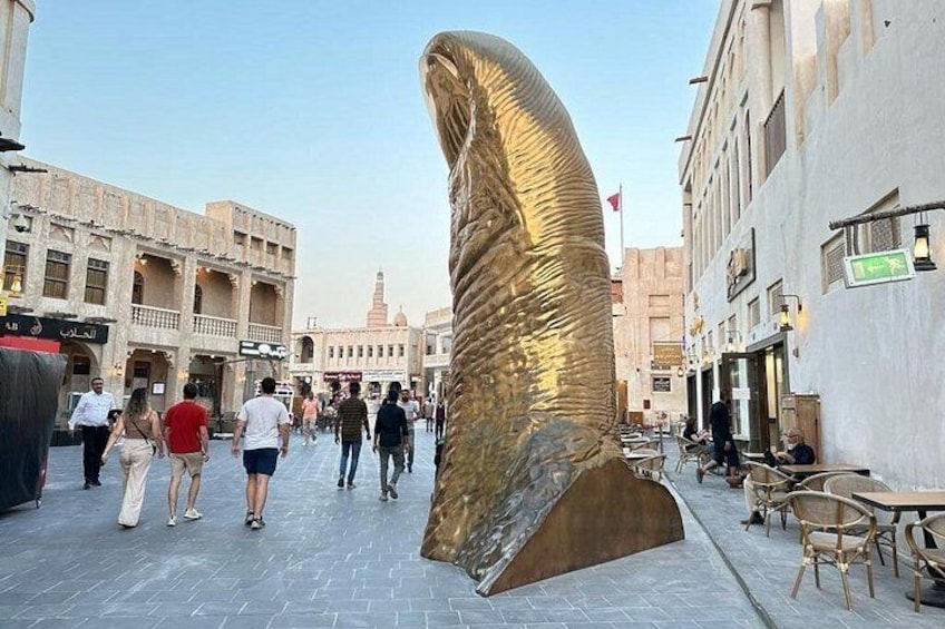 A public square with a large golden spiral sculpture, people walking, and traditional buildings. 1