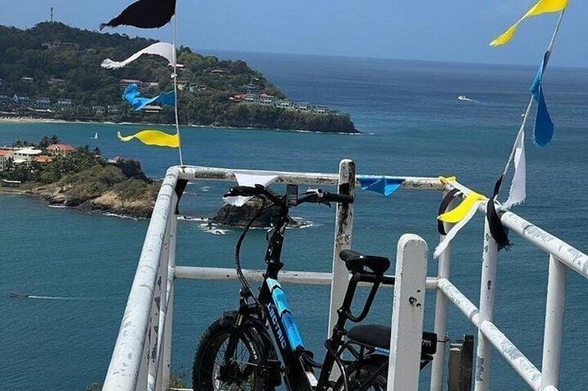 A bicycle parked at a railing overlooking a coastal area with flags and a view of the sea and a distant island. 4