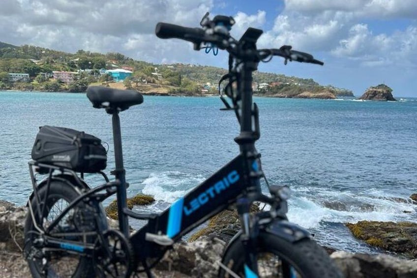 A black and blue electric bike with a basket, parked by a rocky shore with a view of the sea and distant hills. 5