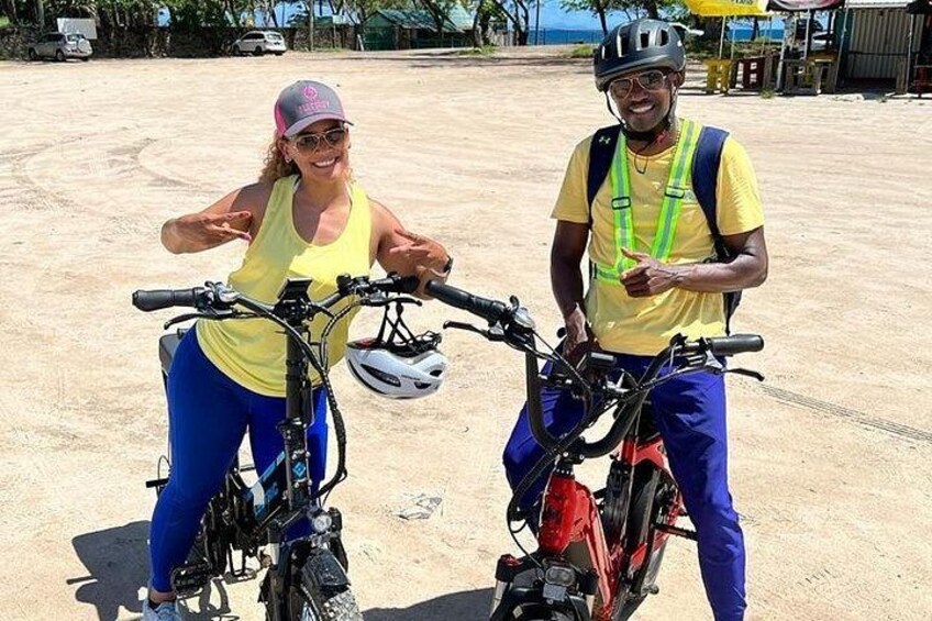 Two people on bicycles in an open area with a dirt ground, a clear sky, and some distant structures. 1