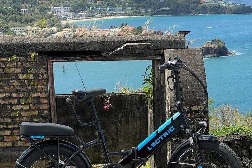 A black electric bike parked in front of a stone wall with a view of the ocean and a rocky island. 2