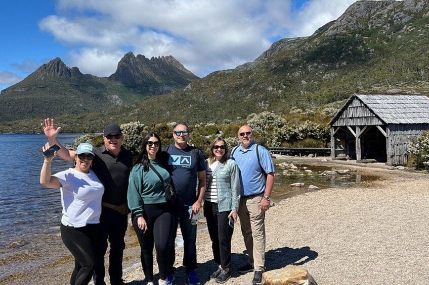 A group of people standing by a lake with mountains in the background and a wooden structure on the shore. 3