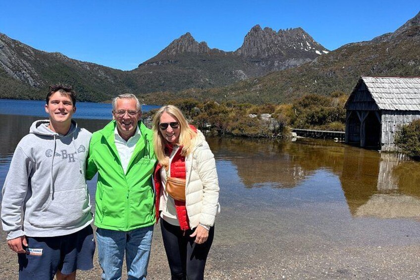 Three people standing by a lake with mountains in the background and a wooden structure on the right. 5