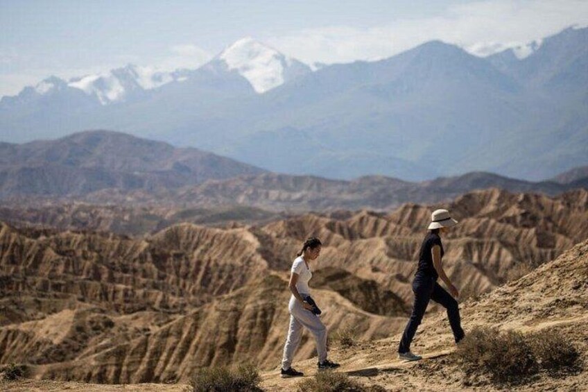 Two people walking on a dry, hilly terrain with a mountainous backdrop. 3
